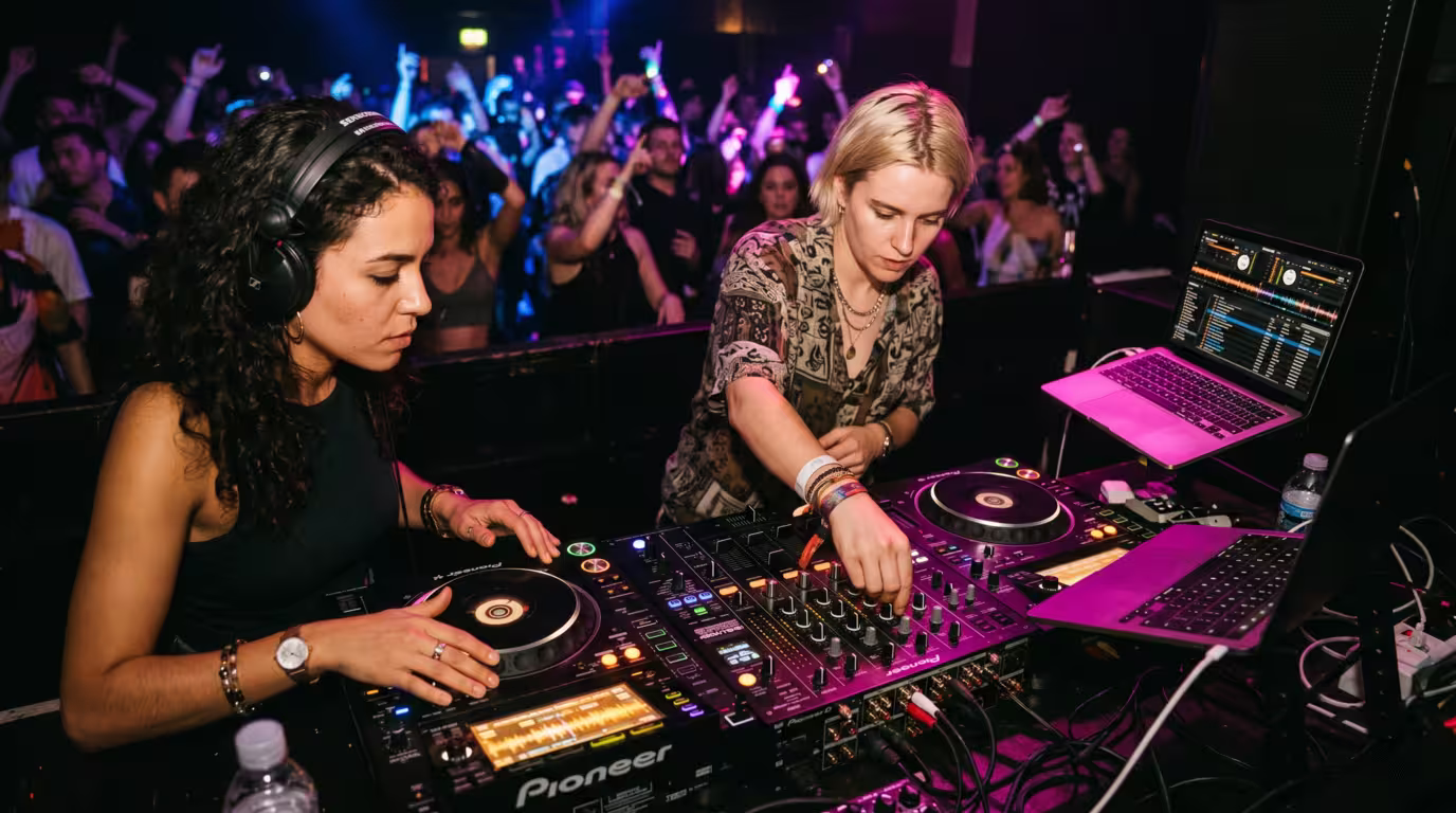 Two DJs sharing a booth during a back-to-back set, one cueing on headphones